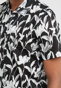 Kortärmad skjorta med knappar i svartvitt tyg, med ett djärvt, abstrakt lövmönster med kontrasterande texturer och släta knappar.