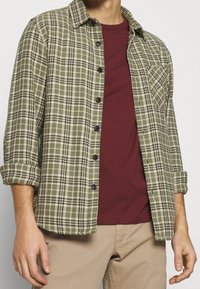 Camicia di flanella a quadri verdi con accenti neri e crema, design a bottoni e maniche arrotolate, indossata sopra una maglietta a girocollo bordeaux.