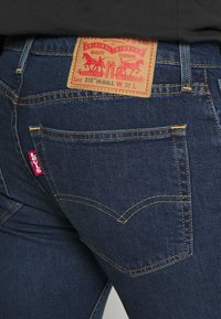 Jeans de mezclilla azul oscuro con un parche de cuero que presenta gráficos en rojo y amarillo, costuras doradas y una pequeña etiqueta roja en el bolsillo trasero.