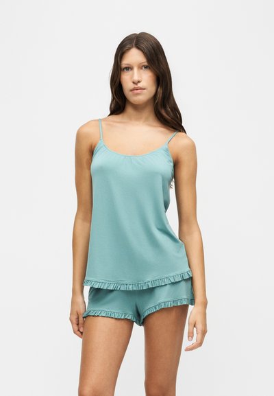 Femme portant un haut sans manches bleu clair et un short assorti avec un bord à volants sur fond uni clair.