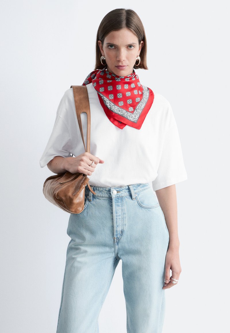 Femme portant une chemise blanche oversize, un jean bleu clair, une écharpe rouge à motifs, des boucles d'oreilles anneaux, tenant un sac à bandoulière en cuir marron.
