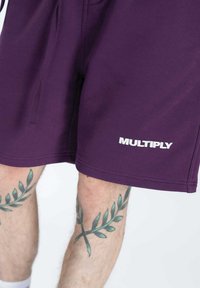Shorts en coton violet avec une taille à cordon, arborant un logo "MULTIPLY" blanc en bas. Texture douce et coupe décontractée.