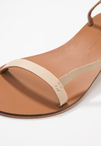 Beige lädertoffel med platt brun sula, med en minimalistisk design och subtila sömnadsdetaljer. Mjuk textur, utan utsmyckningar.