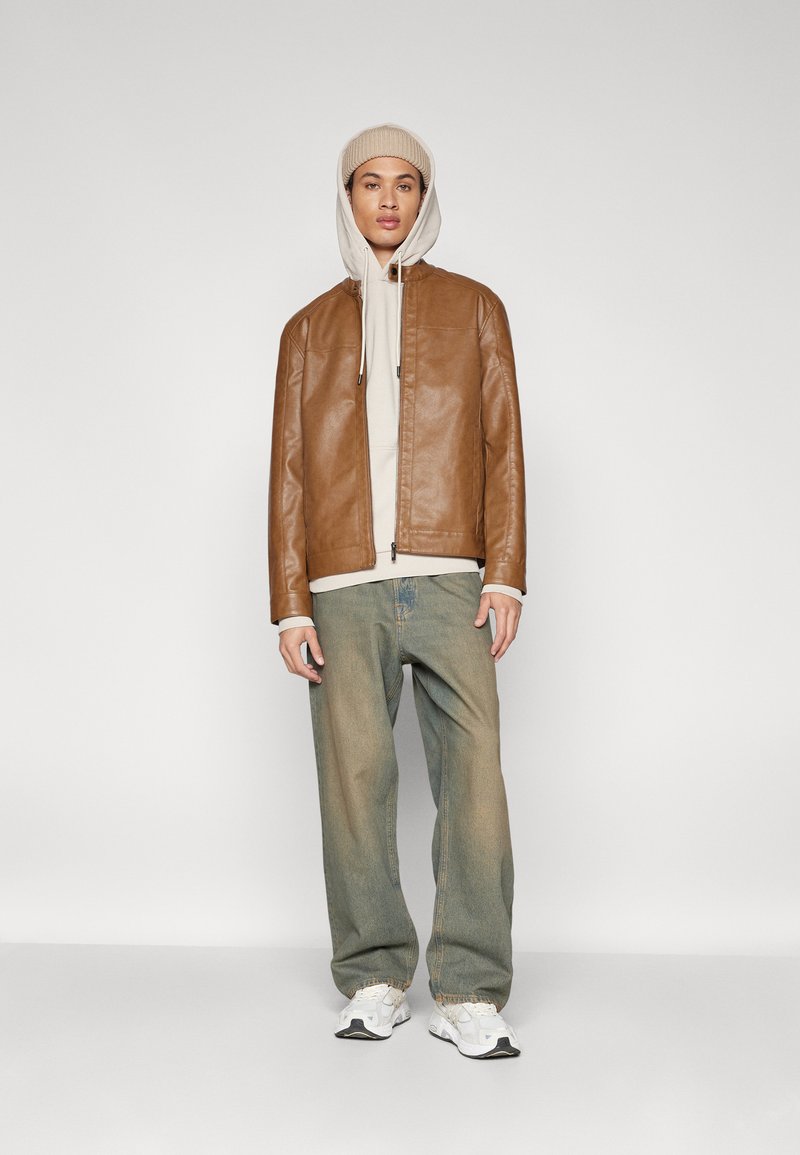 Chaqueta de cuero marrón, sudadera beige, jeans ligeros azul claro relajados y zapatillas blancas. El modelo lleva un gorro de punto beige. Fondo neutro.