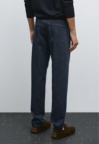 Mörkblå denimjeans med avslappnad passform, femficksdesign och synliga sömnadsdetaljer. Bärs med bruna mocka slip-on-skor.