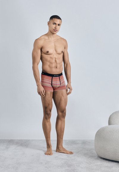 Homme musclé debout pieds nus sur un tapis, portant des caleçons à rayures multicolores, mur gris clair et chaise blanche courbée en arrière-plan.