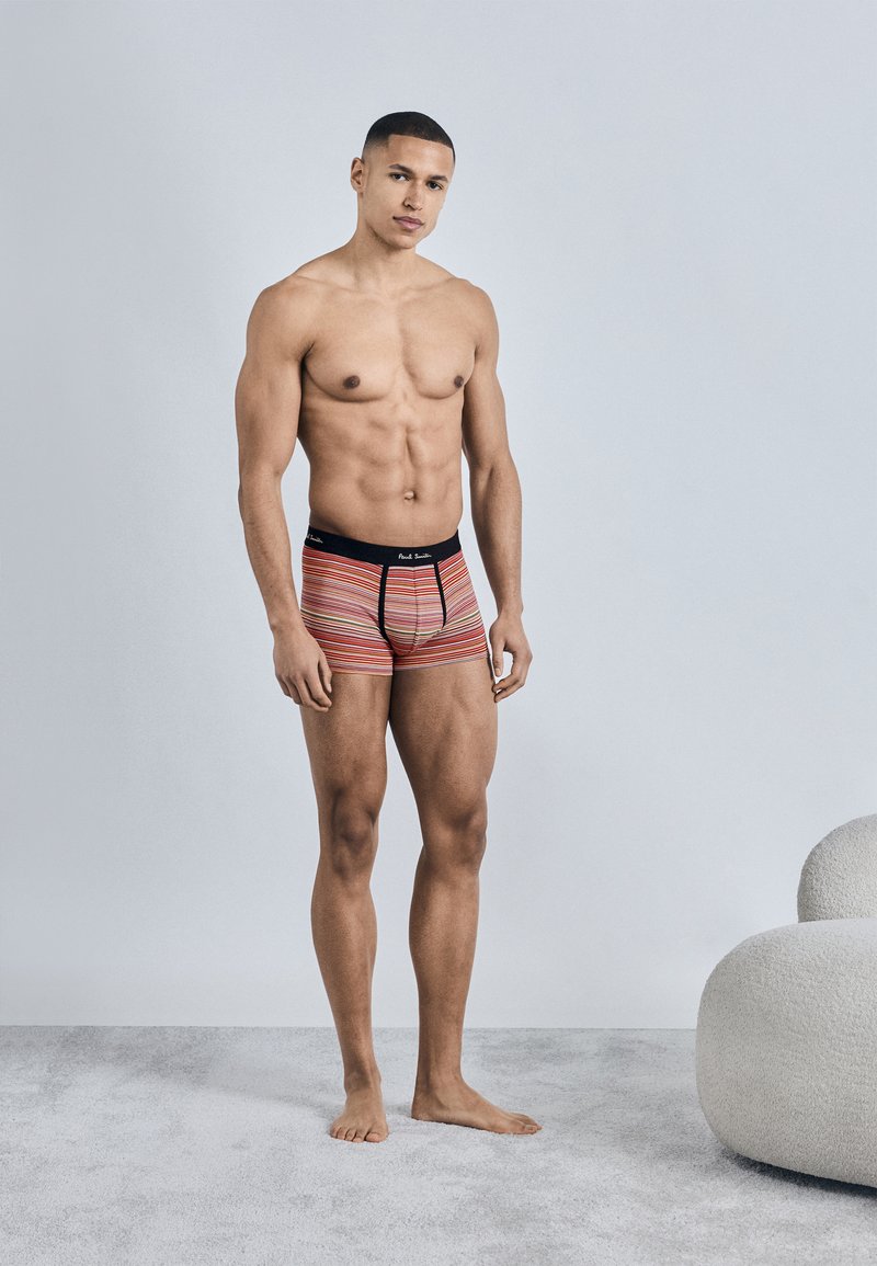 Homme musclé debout pieds nus sur un tapis, portant des caleçons à rayures multicolores, mur gris clair et chaise blanche courbée en arrière-plan.