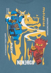 Blå og røde ninja-figurer på en teal skjorte, med gule aksenter og tekst som sier "Ninjago" og "Sterkere sammen, drager reiser seg."