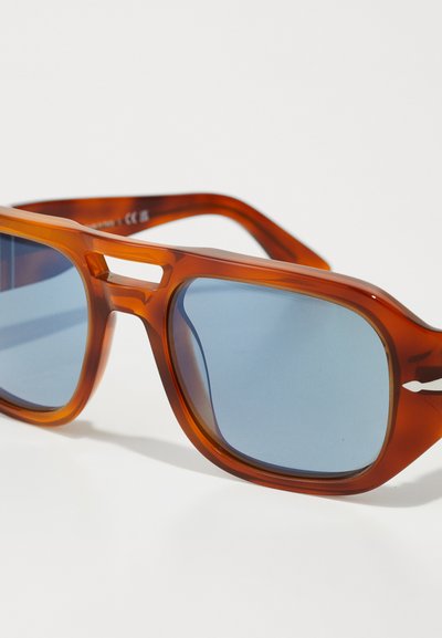 Ochelari de soare din plastic maro cu lentile albastre, design unghiular, rame groase și un detaliu decupat pe punte, oferind un aspect modern.
