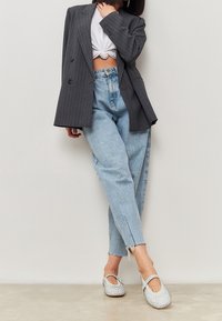 Kobieta w szarym żakiecie w prążki, białym węzłowym topie cropped, jasnoniebieskich spodniach typu high-waist oraz białych balerinach z ćwiekami.