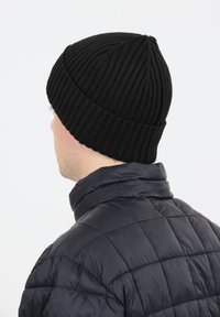 Svart stickad mössa med ribbat mönster, spets framtill och tight passform. Bärs med en svart vadderad jacka med hög krage.