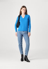 Pulover albastru cu decolteu în formă de V, realizat dintr-un material moale, asortat cu blugi skinny albastre deschise și cizme negre până la gleznă, purtând o geantă neagră texturată.