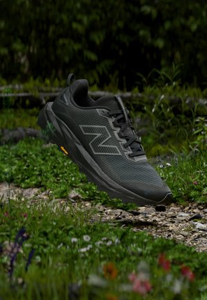 Sort New Balance løbesko med Vibram-sål hævet over en stenrig, græsbevokset udendørs sti med vilde blomster og løv.