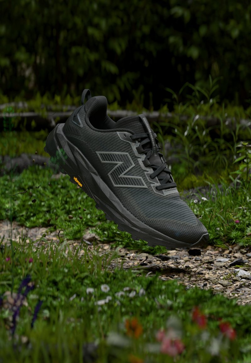 Μαύρο αθλητικό παπούτσι New Balance με σόλα Vibram στον αέρα πάνω από πετρώδη, χορταριασμένη εξωτερική διαδρομή με αγριολούλουδα και φυλλώματα.