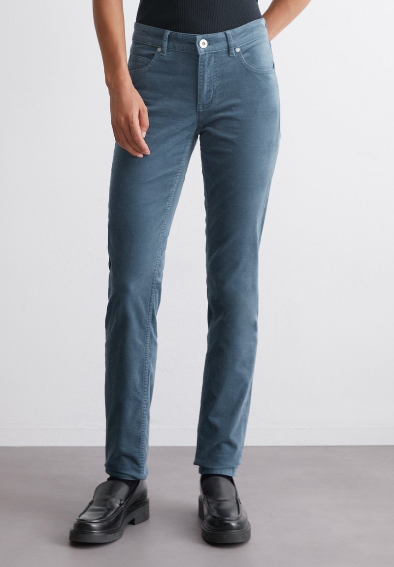 Personne portant un pantalon bleu slim et des mocassins noirs, se tenant devant un fond gris clair uni.