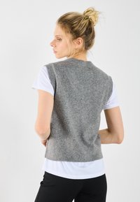 Grå stickad väst med rund halsringning och ärmlös design, bärs över en vit T-shirt, med ribbad nederkant och mjuk textur.