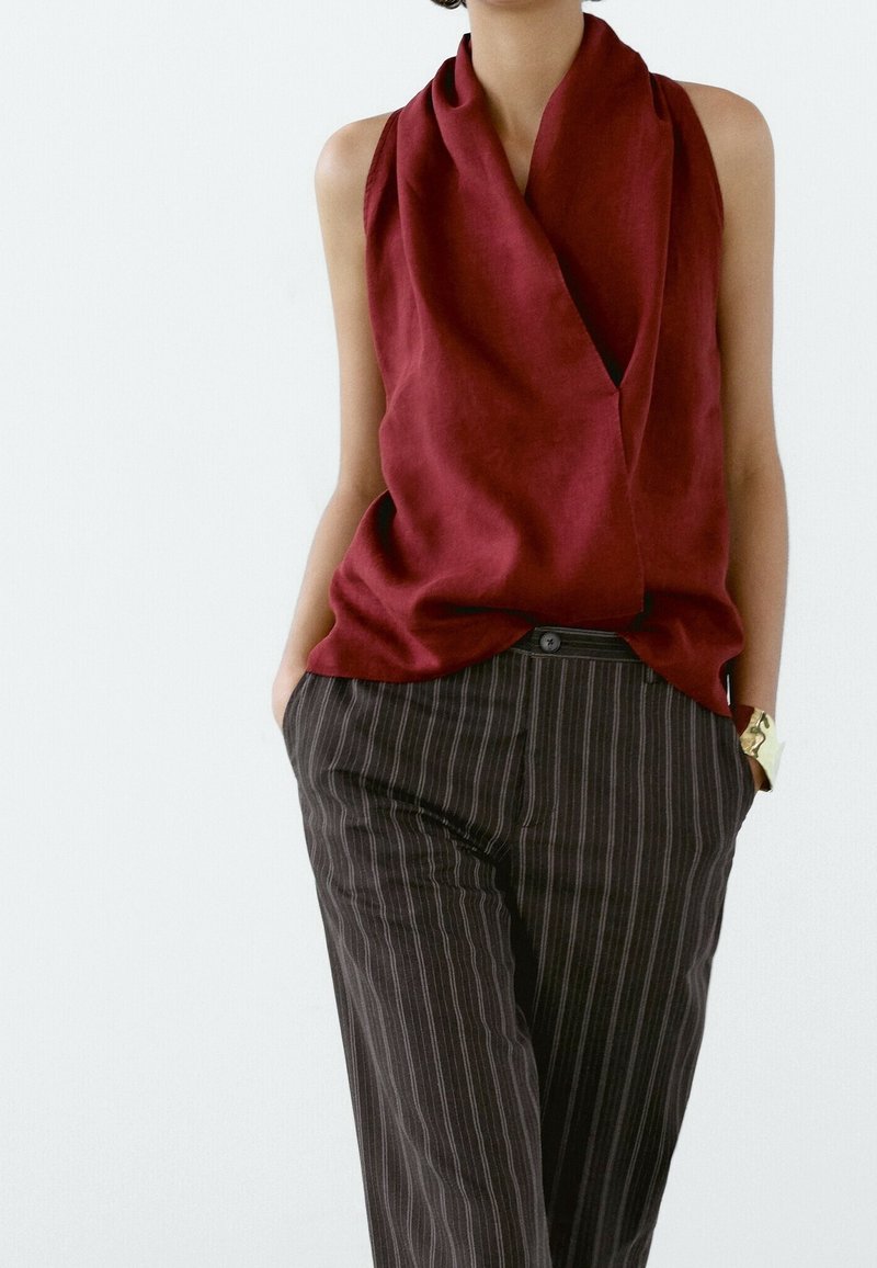 Femme portant un haut sans manches rouge profond drapé et un pantalon à rayures noires, mains dans les poches, avec un bracelet en or au poignet gauche.