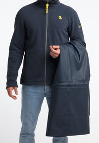Veste polaire marine avec un col montant, une fermeture éclair et un tire-zip jaune. La personne tient un imperméable marine assorti.