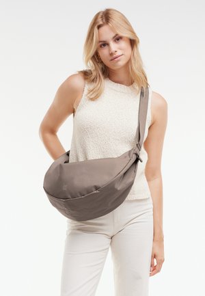 Mujer joven con cabello rubio vestida con una blusa sin mangas blanca y pantalones blancos, llevando un bolso gris grande de tipo bandolera en el hombro.