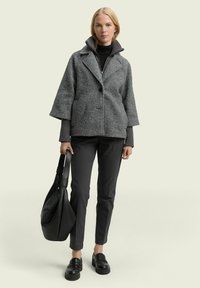 Grijze textuurjack met een kraag, zwart coltruitje, donkere broek en zwarte leren instappers. Houdt een grote zwarte tas vast.