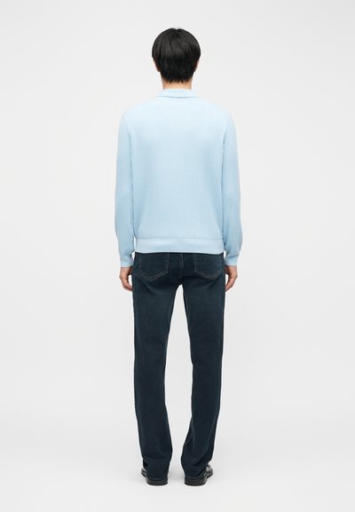 Personne de dos portant un pull côtelé bleu clair, un jean foncé et des chaussures noires sur un fond blanc uni.