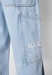 Ljusblå denimbyxor i närbild som visar sidofickan med vit broderad text "KAN... THE ORIGINAL EST. 1989 RDC TRADEMARK".