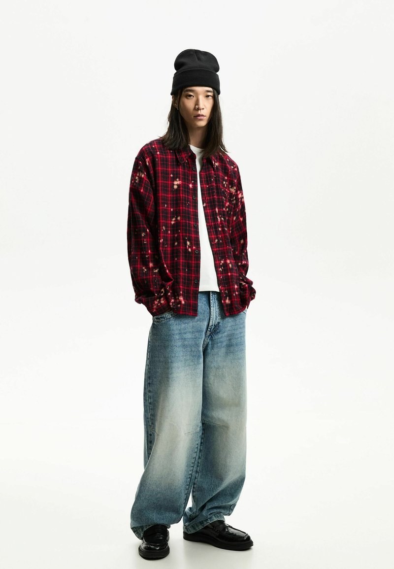 Une jeune personne portant un bonnet noir, une chemise à carreaux rouge décolorée, un t-shirt blanc, un jean bleu clair ample et des chaussures noires, se tenant contre un fond blanc.