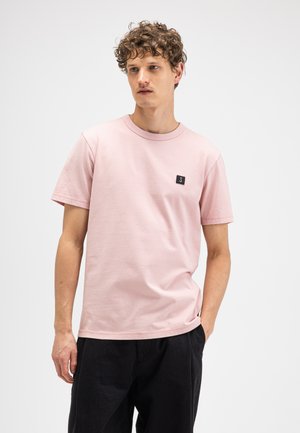 Young man with curly hair wearing a light pink short-sleeve t-shirt with a small black patch and black pants, standing against a plain background.