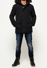 Svart parka med faux fur huva, knäppning med knappar och två framfickor. Bärs över trasiga blå jeans och svarta snörkängor.
