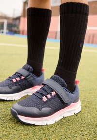 Calzini neri fino al ginocchio abbinati a scarpe sportive blu navy e rosa con tomaia in mesh, cinturino in Velcro e suola testurizzata su superficie erbosa.