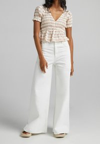 Jeans bianchi a gamba larga abbinati a un top elasticizzato a righe beige e rosa con maniche a palloncino corte e scollatura a V. Texture liscia.