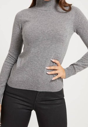 Frau trägt eng anliegenden grauen Rollkragenpullover und schwarze Hose, rechte Hand ruht auf der Hüfte vor schlichtem Hintergrund.