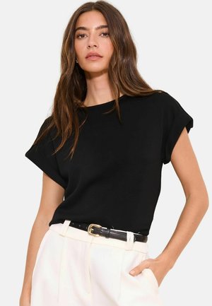 Žena s dlhými hnedými vlasmi, oblečená v čiernom krátkym rukávom tričku zastrčenom do bielych nohavíc s čiernym opaskom, rukou v vrecku, neutrálne pozadie.