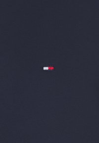 Logo rectangular pequeño con secciones en blanco, rojo y azul bordadas sobre un fondo de tela ribeteada de color azul oscuro.