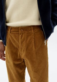 Braune Cordhose mit vertikaler Rippstruktur, ausgestattet mit einem Knopfverschluss und geplättetem Design, kombiniert mit einem leichten Pullover und einer marineblauen Jacke.