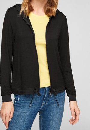 Kvinde iført en sort lynlås-hættetrøje over en gul skjorte og blå jeans, stående med hænderne afslappet ved siderne.