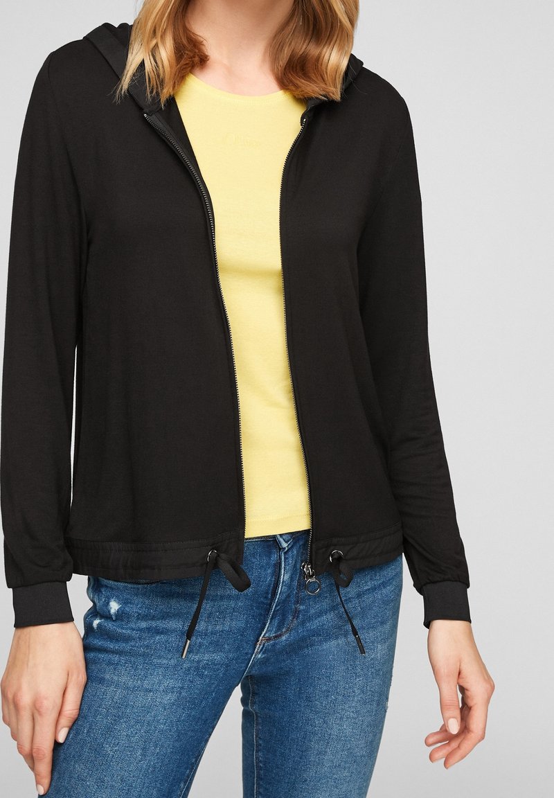 Femme portant un sweat à capuche noir zippé sur un t-shirt jaune et un jean bleu, debout avec les mains détendues le long du corps.