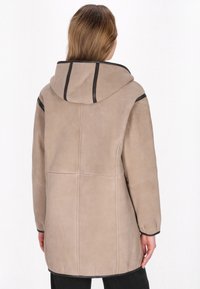 Abrigo de ante beige con capucha, que presenta detalles en negro a lo largo de las costuras, un corte relajado y costuras visibles en la parte posterior.
