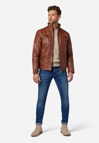 Bruine leren jas met opstaand kraagje, ritszakken en een zachte textuur, gecombineerd met lichtblauwe jeans en beige enkellaarsjes.