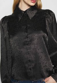 Musta satiinipaita kukkakuviolla, jossa on nappikiinnitys edessä, iso kaulus ja puffy-hihat. Sileä pinta kiiltävällä viimeistelyllä.