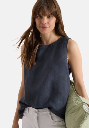 Jeune femme aux longs cheveux bruns portant un haut bleu foncé sans manches et un pantalon clair, tenant une veste matelassée vert clair.