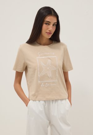 Femme aux longs cheveux foncés portant un t-shirt beige avec un motif d'étoile de mer blanche et un texte en français, associée à un pantalon blanc ample, mains dans les poches.