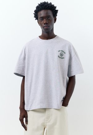 Giovane uomo con capelli corti ricci che indossa una maglietta oversize grigio chiaro con la scritta "Los Angeles California" e pantaloni beige, in piedi su sfondo bianco.