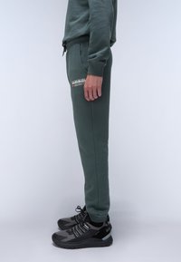 Grüne Sweatpants mit elastischem Bund, seitlichen Taschen und Logo-Detail. Kombiniert mit schwarzen Sneakers mit hellgrauen Akzenten und Schnürsenkeln.