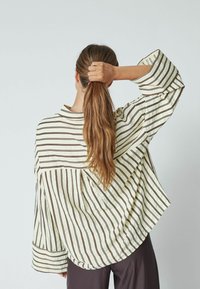 Chemise à rayures jaune clair avec des rayures verticales marron, au design oversize, avec des manches larges et une coupe ample, portée avec un pantalon sombre.
