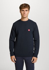 Marineblå strikkegenser med rund hals, ribbestrikkede mansjetter og kant, med en liten rød logo på brystet. Materialet ser teksturert og varmt ut.