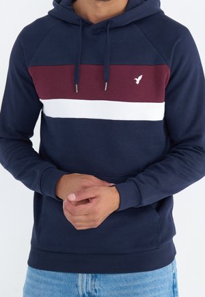 Man draagt een marineblauwe hoodie met bordeauxrode en witte horizontale strepen en een klein vogel-logo, handen in elkaar gevouwen, gecombineerd met lichtblauwe spijkerbroek.