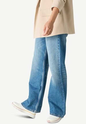 Osoba ubrana w jasnobeżową marynarkę i szerokie niebieskie dżinsy z białymi trampkami, stojąca na jednolitym tle.