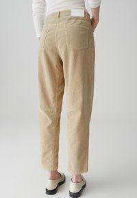 Pantaloni in corduroy beige con taglio dritto, dotati di tasche posteriori e una trama sottile. La vita presenta un'etichetta bianca. Scarpe nere completano il look.