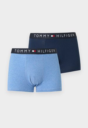 Mężczyzna bez koszulki, stojący boso, ubrany w jasnoniebieskie bokserki Tommy Hilfiger, patrzący w bok na jednolitym białym tle.
